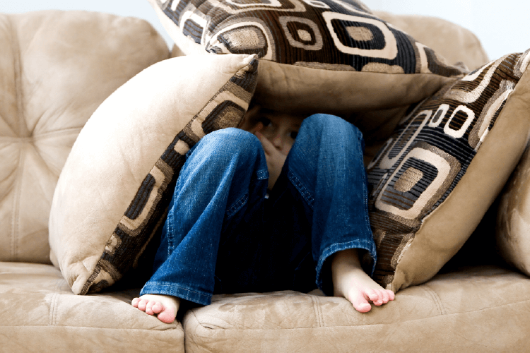 little boy hiding under beige couch pillows